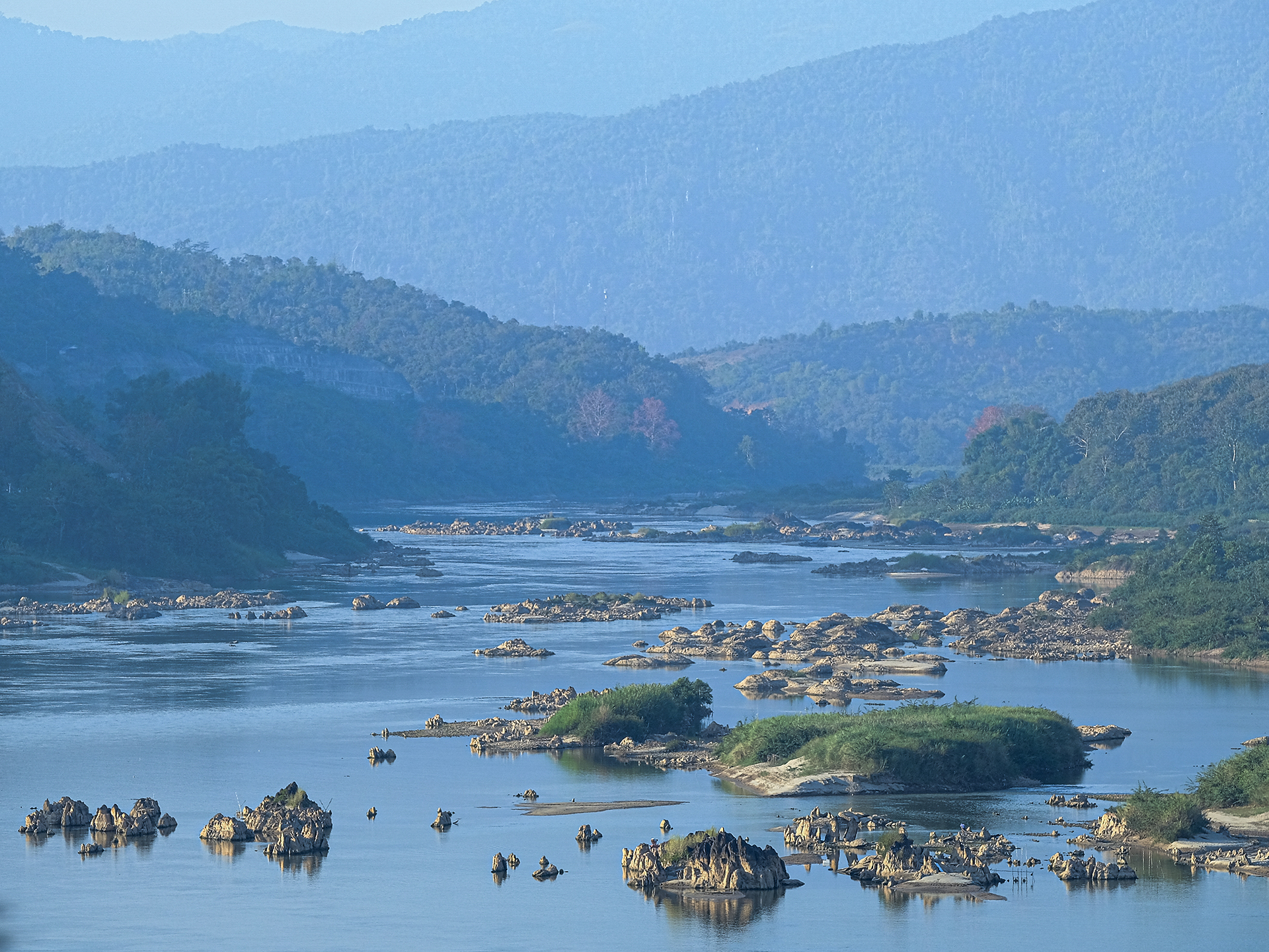 Mekong River with small grassy islands dispersed throughout the river with mountainous hills in background.