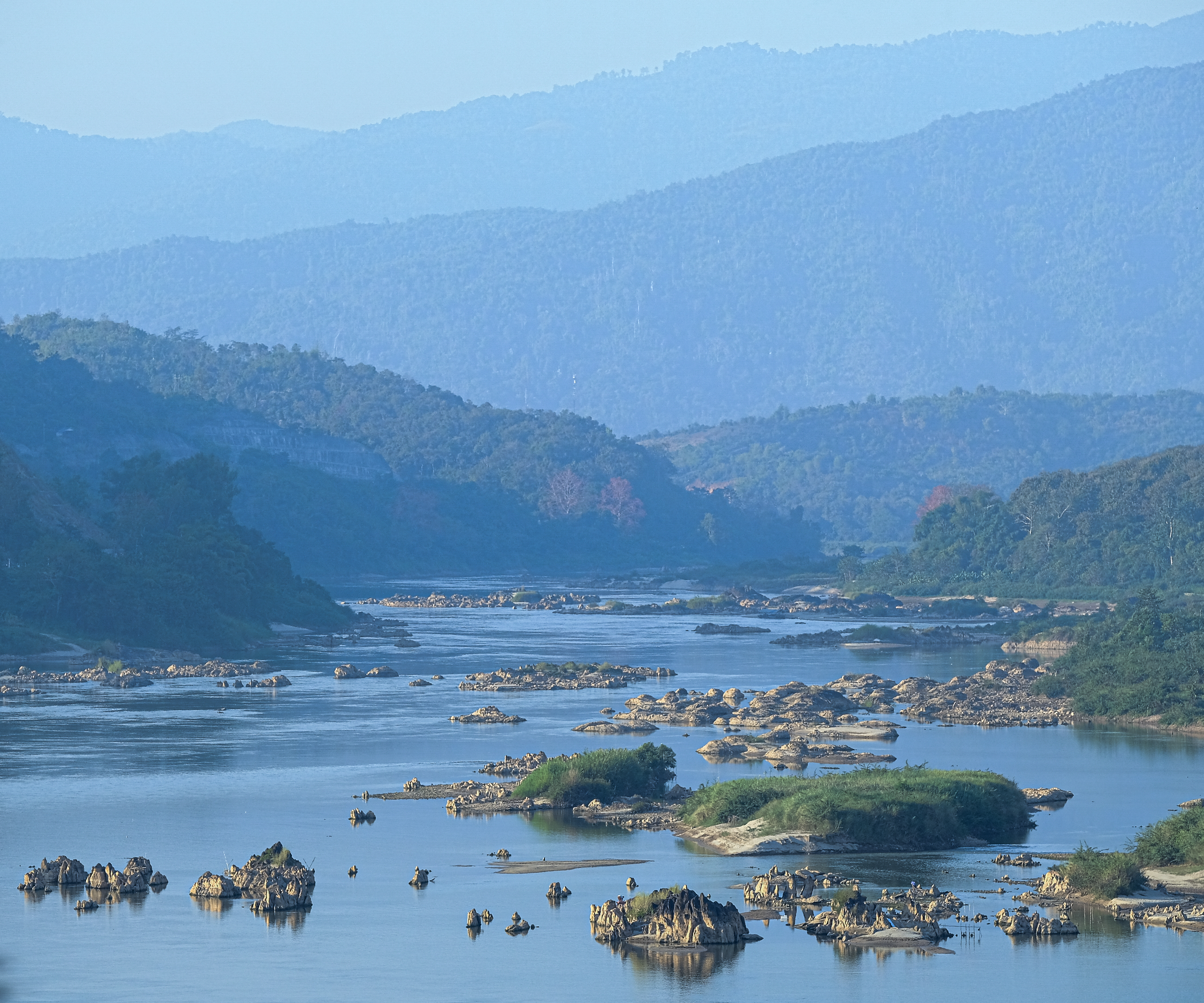 Mekong River with small grassy islands dispersed throughout the river with mountainous hills in background.