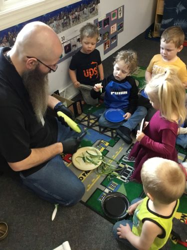 Head Start teacher sits on the floor with a group of young children, showing them how to prepare cucumbers. The children watch closely and hold small plates as they participate in a lesson on healthy eating.