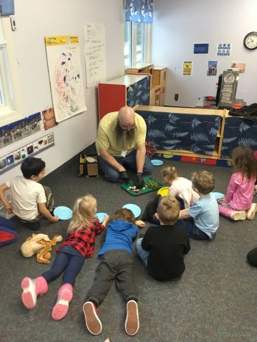 A Head Start teacher sits on the floor cutting radishes with young students as they prepare for a lesson on healthy eating. The children sit ready to taste test the vegetables with plates in hand.