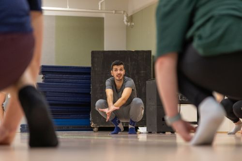 A dance instructor crouches while MSU students in a circle around him mirror the pose.