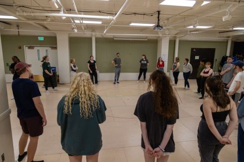 MSU students stand in a wide circle in a rehearsal studio listening to a dance instructor who is wearing a black shirt and gray athletic pants.