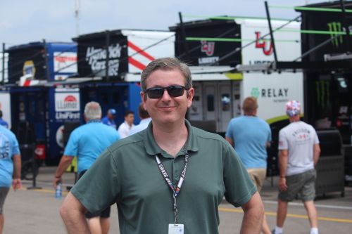 David Ferguson wearing sunglasses stands in a NASCAR Cup Series garage, with teams and fans in the background.