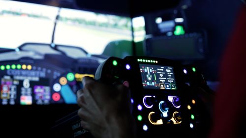 Ricky Taylor holds racing simulator steering wheel while racing a virtual track, with colorful lights representing real elements in a race car.