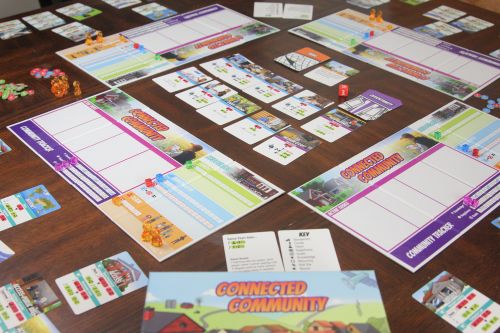 Photo of the Connected Community board game on a table, showing a town‑themed board with buildings, streets, game tokens, and cards used for community decision‑making.