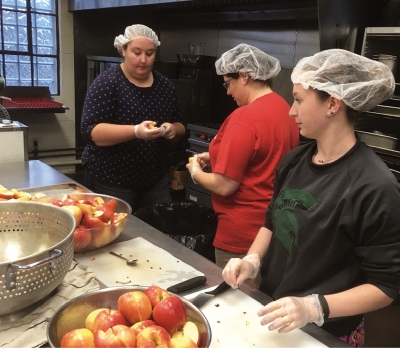 Brittany Motz, Mara Stein, and Emily DiGiovanni prepare a meal for delivery. This menu has baked apples as the fruit/dessert portion of the meal.